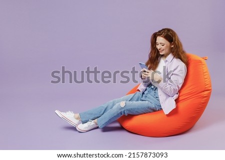 Full size body length young redhead curly woman 20s wear white T-shirt violet jacket sit in bag chair hold in hand use mobile cell phone isolated on pastel purple color wall background studio portrait