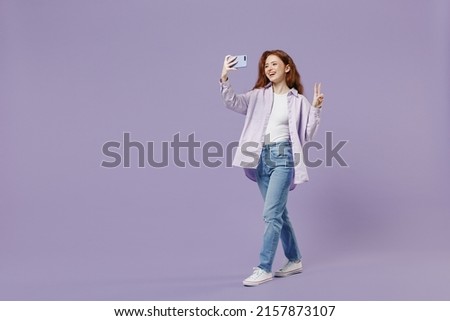 Full size body length young redhead woman 20s wears white T-shirt violet jacket do selfie shot on mobile cell phone show victory sign isolated on pastel purple color wall background studio portrait