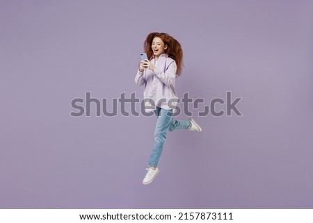 Full size body length young redhead curly green-eyed woman 20s wears white T-shirt violet jacket jump hold in hand use mobile cell phone isolated on pastel purple color wall background studio portrait Royalty-Free Stock Photo #2157873111