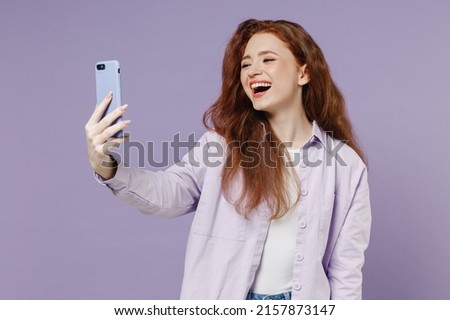 Smiling overjoyed fun young redhead curly green-eyed woman 20s wears white T-shirt violet jacket doing selfie shot on mobile cell phone isolated on pastel purple color wall background studio portrait.