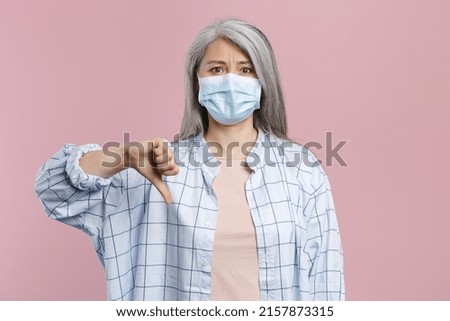 Gray-haired asian woman in white shirt sterile face mask to safe from coronavirus virus covid-19 during pandemic quarantine showing thumb down isolated on pastel pink background, studio portrait