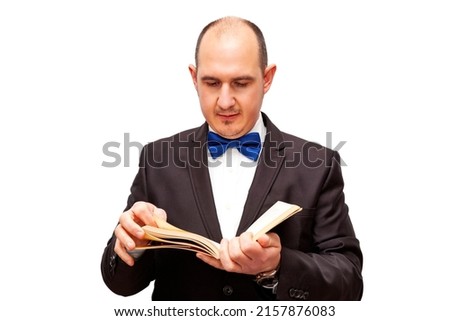 A bald Caucasian adult male dressed in a black suit, white shirt and blue bow tie is reading a book. The background is white.