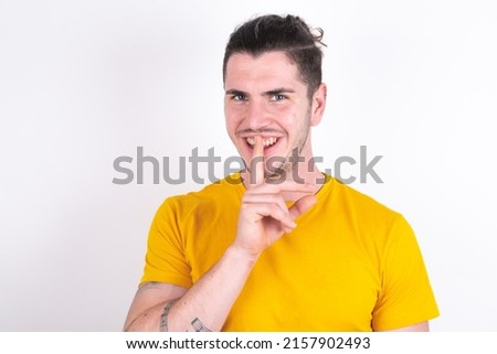 Smiling Young caucasian man wearing yellow t-shirt over white background makes shush gesture, holds fore finger over lips hides secret. Be mute, please.