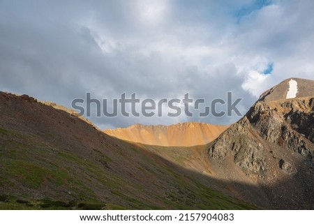 Dramatic alpine landscape with sunlit wide sharp mountain ridge under overcast sky at changeable weather. Atmospheric mountain scenery with large sharp rocks on ridge top in sunlight under cloudy sky. Royalty-Free Stock Photo #2157904083