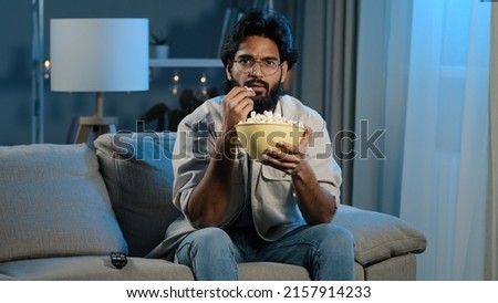 Serious interested Arabic Hispanic Indian bearded man guy wearing glasses 30s male with popcorn watching TV at home sofa late evening night relaxing enjoying movie online sport game attentively watch Royalty-Free Stock Photo #2157914233
