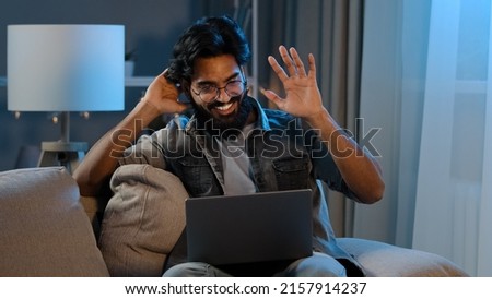 Arabic indian bearded man millennial guy businessman freelancer sitting at home sofa in evening night dark late time waving hello in webcam online video call conference with laptop remote conversation Royalty-Free Stock Photo #2157914237