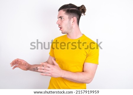 Displeased Young caucasian man wearing yellow t-shirt over white background keeps hands towards empty space and asks not come closer sees something unpleasant