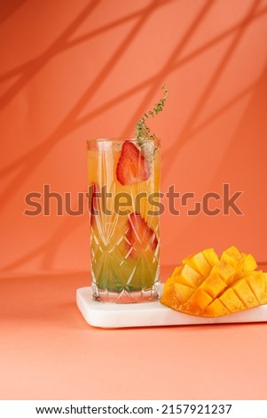 tall cocktail glass filled with yellow juice and fresh strawberries and cut in cubes mango half on tropical orange background, gobo mask effect