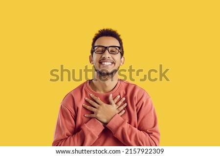 Happy black man in glasses smiling and holding his hands on his chest to express endless gratitude and appreciation. Cheerful ethnic guy says thank you from the bottom of his heart for gift or present Royalty-Free Stock Photo #2157922309