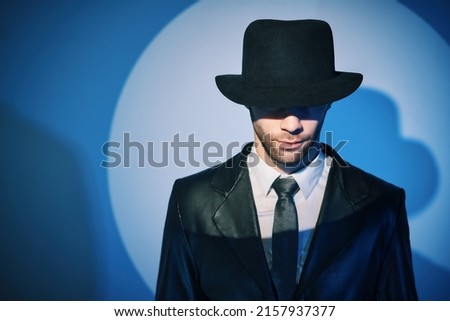 Portrait of handsome man in black coat hiding face behind his hat in the spotlight on studio background. noir film style. Private detective, investigation, secret concept. 