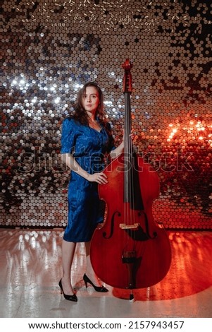 Girl musician with long hair in blue dress poses with the double bass against the glitter mosaic background.