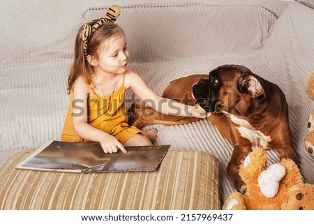 a little girl in a yellow dress and wavy hair reads a book and shows pictures to her big red dog on the sofa at home