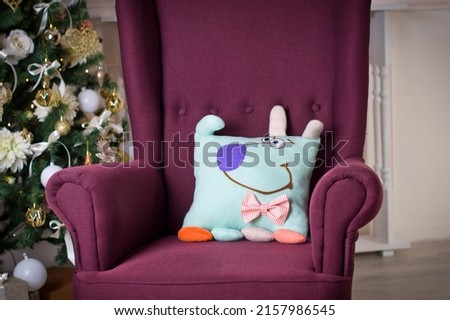 Soft funny handmade colorful pillow in the form of a dog stot on a burgundy chair against the backdrop of a Christmas tree and gifts