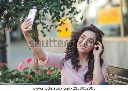 Young curly woman uses a phone for social networks. A model takes a selfie on the street of a European city in summer.