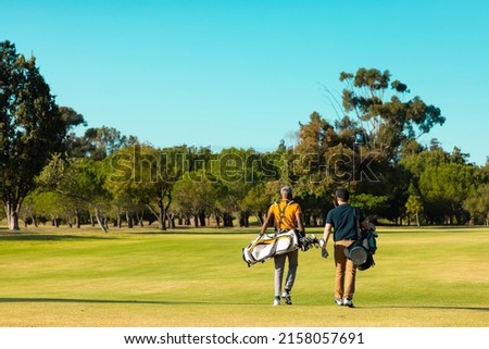 Rear view of multiracial young male friends with golf bags walking at golf course against clear sky. copy space, summer, golf, unaltered, friendship, togetherness, nature, sport, weekend activities. Royalty-Free Stock Photo #2158057691