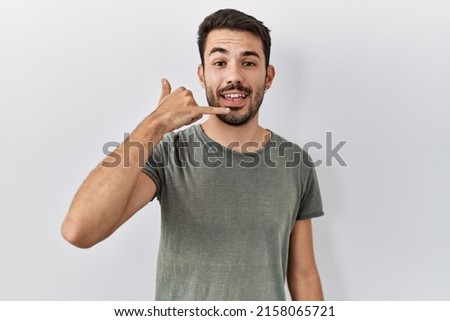 Young hispanic man with beard wearing casual t shirt over white background smiling doing phone gesture with hand and fingers like talking on the telephone. communicating concepts. 