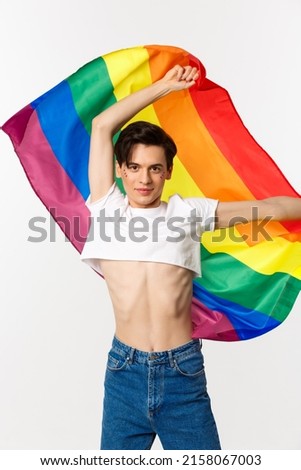 Vertical view of happy queer person in crop top and jeans waving raised rainbow flag, celebrating lgbtq holiday, standing over white background