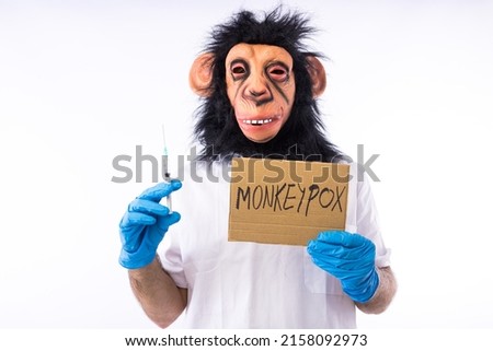 Person dressed in a monkey with a mask, with a medical nurse's suit, with a sign that reads: 'MONKEYPOX', and a syringe, on a white background. Pandemic, virus, epidemic, Nigeria and smallpox concept. Royalty-Free Stock Photo #2158092973