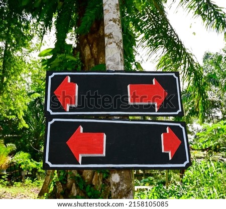 Two black wooden signs drawn with a white border and red arrows of different sizes pointing in opposite directions. Installed side by side on the garden plants. Blank labels are used to enter text.