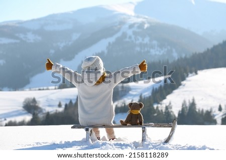 Сhild with toy teddy bear sits on a sled and looks at the winter snowy mountains.Winter family vacantion. Christmas celebration and winter holidays. Winter fun and outdoor activities with kids Royalty-Free Stock Photo #2158118289