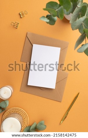 Top view vertical photo of workplace paper sheet over craft paper envelopes gold pen binder clips candles on rattan serving mat and vase with eucalyptus on isolated orange background with blank space