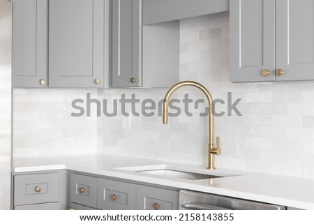 A kitchen sink detail shot with a gold faucet, marble backsplash, grey cabinets, and gold hardware. Royalty-Free Stock Photo #2158143855