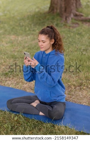 a woman in the park does sports or fitness on a yoga mat. a European woman with curly hair in a blue hoodie is chatting on a smartphone after a fitness class. a happy woman is relaxing in the park