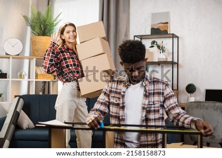 Cute caucasian woman making peace sign joyfully to camera while holding cardboard boxes at room. African american man with tape measuring modern shelves. New apartment for young family.