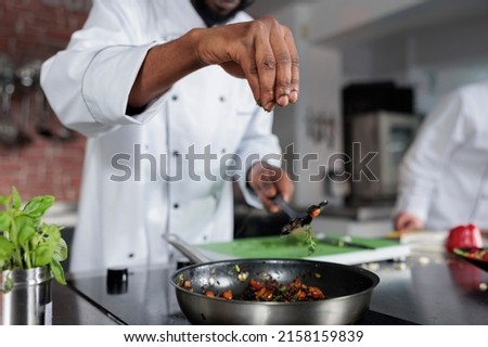Master chef seasoning dish prepared for food contest held at fine dining restaurant. Head cook throwing fresh chopped herbs in pan to improve taste of meal while in professional kitchen. Royalty-Free Stock Photo #2158159839