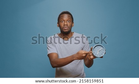 Stressed man checking time on wall clock and running late to work, looking at hour and minutes. Young adult in delay, doing alarm countdown to measure time, being impatient and punctual. Royalty-Free Stock Photo #2158159851