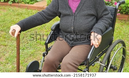 Elderly woman resting and sunbathing in the garden, in a wheelchair and holding a wooden cane.