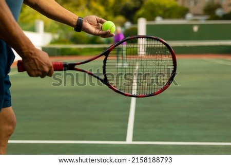 Midsection of biracial senior man holding racket serving tennis ball while playing at tennis court. unaltered, sport, competition, retirement, healthy and active lifestyle concept. Royalty-Free Stock Photo #2158188793