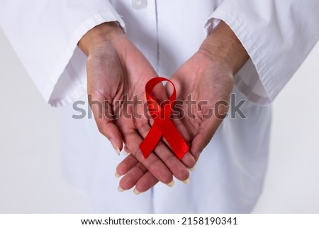 Midsection of african american mid adult female doctor's hands with red aids awareness ribbon. doctor, hiv, aids, red, disease, ribbon, awareness, support, healthcare, hospital, medical occupation. Royalty-Free Stock Photo #2158190341