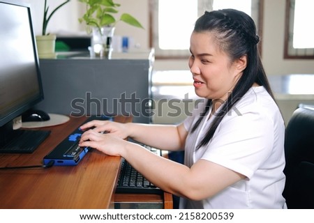 Asian woman with blindness disability using computer with refreshable braille display or braille terminal a technology assistive device for persons with visual impairment in workplace. Royalty-Free Stock Photo #2158200539