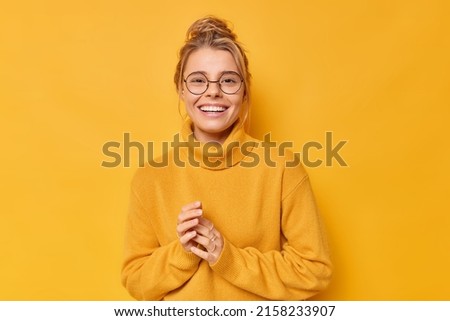 Pretty blonde European woman smiles toothily keeps hands together feels satisfied wears round spectacles and casual loose jumper isolated over vivid yellow background. Happy emotions concept Royalty-Free Stock Photo #2158233907