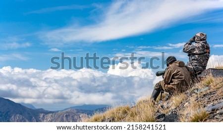Two men in protective camouflage clothing on trophy hunt in the mountains. Hunter and the huntsman are searching in optical devices. Royalty-Free Stock Photo #2158241521