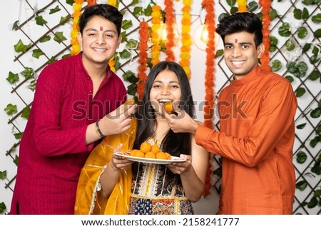 Portrait of happy indian young kids or bothers and sister wearing traditional cloths having fun eating laddu or laddoo sweets celebrating festival  diwali or rakshabandhan, flower decoration. Royalty-Free Stock Photo #2158241777