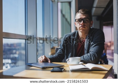 Portrait of a man, IT professional, working remotely with a modern laptop, sitting at a table and smiling at the camera during a break, a happy human programmer in vision correction glasses