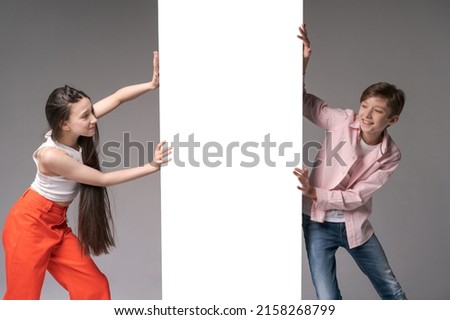 Surprised teenagers are pushing banner with copy space. Portrait cute girl and guy looking at camera against a gray wall studio. Childish fashion and happy emotions concept of caucasian teenagers Royalty-Free Stock Photo #2158268799