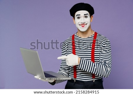 Charismatic smiling fun young mime man with white face mask wears striped shirt beret hold use work point finger on laptop pc computer isolated on plain pastel light violet background studio portrait