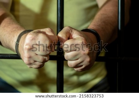A handcuffed man clenches his fists behind bars in a cell. Concept: a prisoner in a courtroom, a court sentence to a convicted person, a prison term 1. Royalty-Free Stock Photo #2158323753