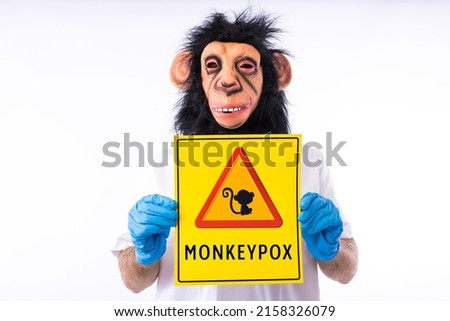 Person dressed in a monkey mask, in a medical nurse's suit, holding a sign with the danger sign and a monkey, which reads: MONKEYPOX, on a white background. Virus, epidemic, Nigeria and smallpox. Royalty-Free Stock Photo #2158326079