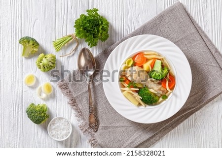 chicken spring soup with broccoli, carrots, parsnip, leek and pasta in white bowl on white wooden table with spoon and ingredients, horizontal view from above, flat lay