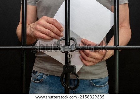 A handcuffed prisoner behind bars holds a white sheet of paper in his hand.  Concept: petition for pardon, court verdict, defense without a lawyer. Royalty-Free Stock Photo #2158329335