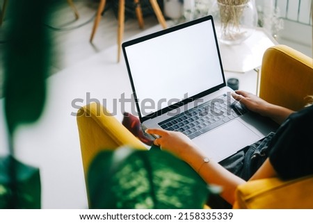 Young caucasian woman sitting on sofa at home and using laptop computer with white blank screen, mock-up.