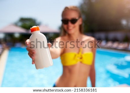 Happy woman shows suntan or sunscreen cream white bottle over the blurred swimming pool background. Tanned slim girl wearing yellow bikini swimwear and holds sun lotion. White bottle with copy space. Royalty-Free Stock Photo #2158336181