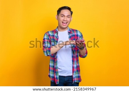 Cheerful young Asian man in plaid shirt holding money banknotes and looking at camera isolated on yellow background