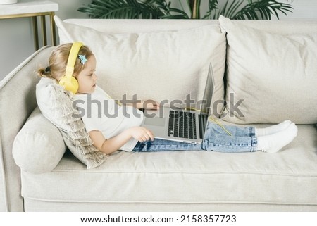 Little girl 4 years old in yellow headphones in front of a laptop. The child looks at the computer screen. The baby watches cartoons while mom is busy. Remote learning for preschool children.