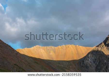 Dramatic alpine landscape with sunlit wide sharp mountain ridge under overcast sky at changeable weather. Atmospheric mountain scenery with large sharp rocks on ridge top in sunlight under cloudy sky. Royalty-Free Stock Photo #2158359535