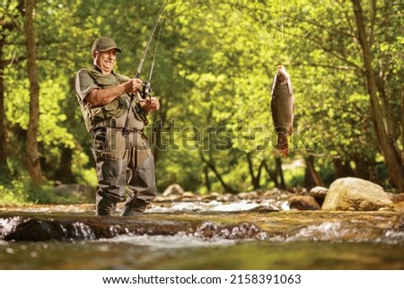 Mature fisherman pulling a carp fish out of a river in the wood Royalty-Free Stock Photo #2158391063